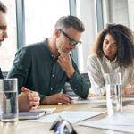 two businessmen and a businesswoman sitting at a desk in an office and reviewing a document